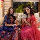 Two women sitting on a couch holding small boxes, with a decorative window and colorful tassels in the background.