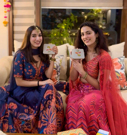 Two women sitting on a couch holding small boxes, with a decorative window and colorful tassels in the background.
